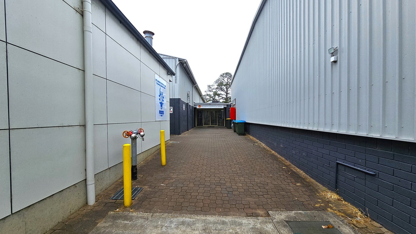 In the middle is an alleyway featuring a brown, brick pathway that is uneven in some areas. The Recreation Centre is on the right, a building with corrugated walls. On the left is another grey building for the Football Club. Yellow bollards are on the left, along with a fire hydrant. At the end is the entrance to Adelaide Hills Recreation Centre.