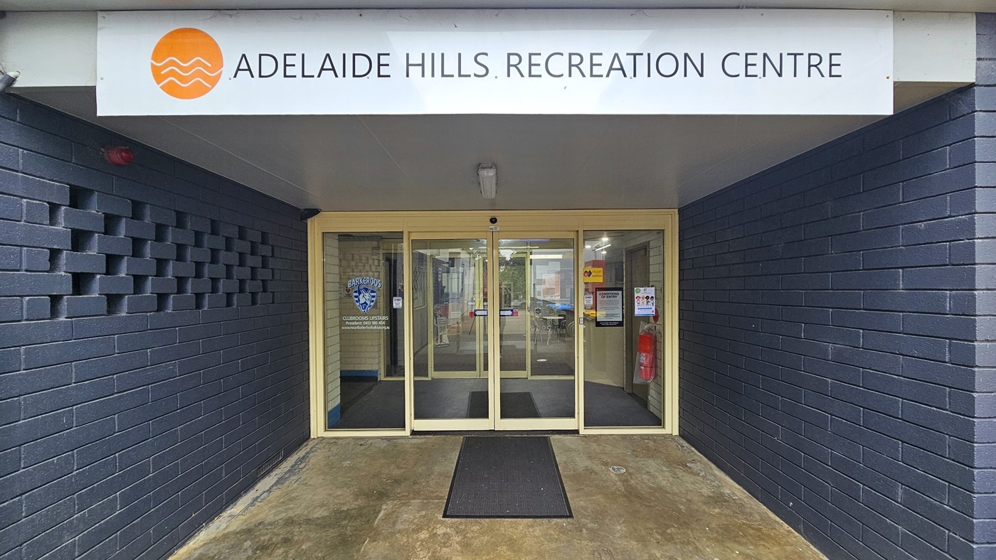The entrance is enclosed by dark blue brick walls. The door consists of two sets of automatic, glass sliding doors with stickers and posters on the wall, including a fire extinguisher. A non-slippery mat is placed on the doorstep. A distinctive sign, 'Adelaide Hills Recreation Centre,' is positioned on top near the ceiling.