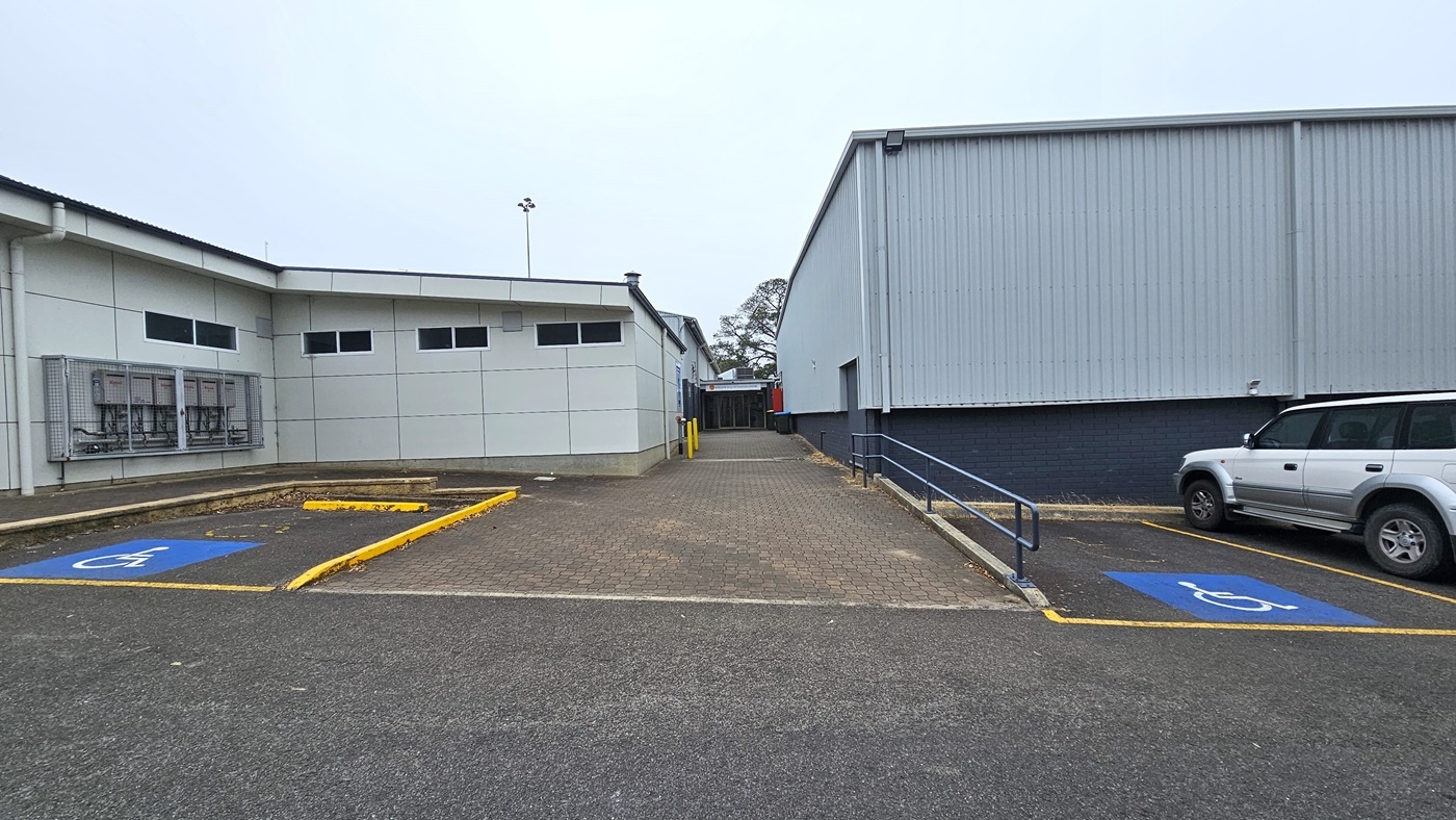 There are two accessible car parks, marked with blue wheelchairs, on the alley's left and right sides. A handrail is installed on the right, and a white car occupies one of the parking spaces. The alley, made of brown bricks, leads to the entrance between the two buildings. The building on the right is constructed with dark blue concrete bricks and corrugated walls, while the building on the left is painted white. An electrical box is affixed to the left wall near the parking.