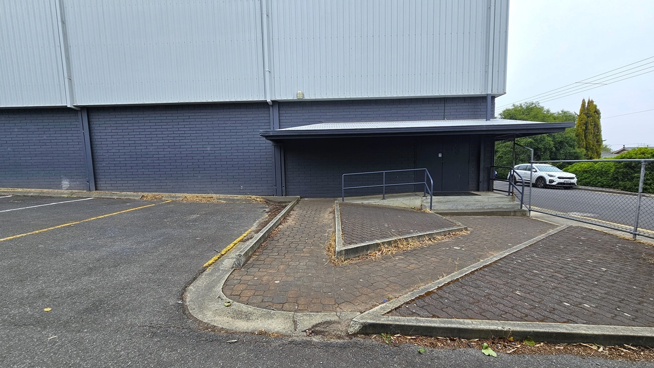 Next to a building made of dark blue bricks and corrugated walls is the parking area. An accessible ramp entrance is on the right, situated on a sloping brick path. To the right, there is a chain fence with a white car parked on the other side.