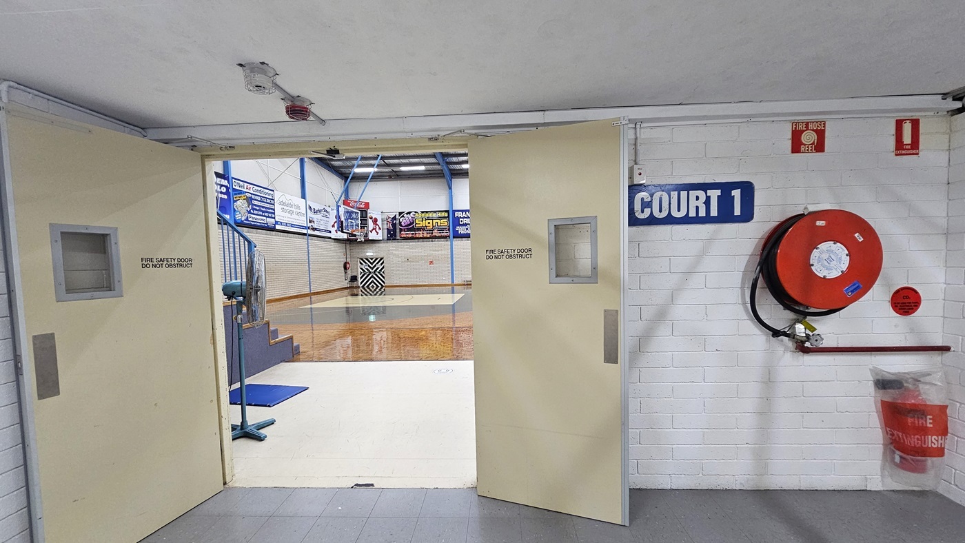 The entrance door, painted in white, is made of wood. To the right, a sign reads "Court 1". On the left wall, a fire extinguisher and a fire hose reel are mounted. Inside, a section of the court is visible, showcasing the timber floor, exposed beams, and signages.