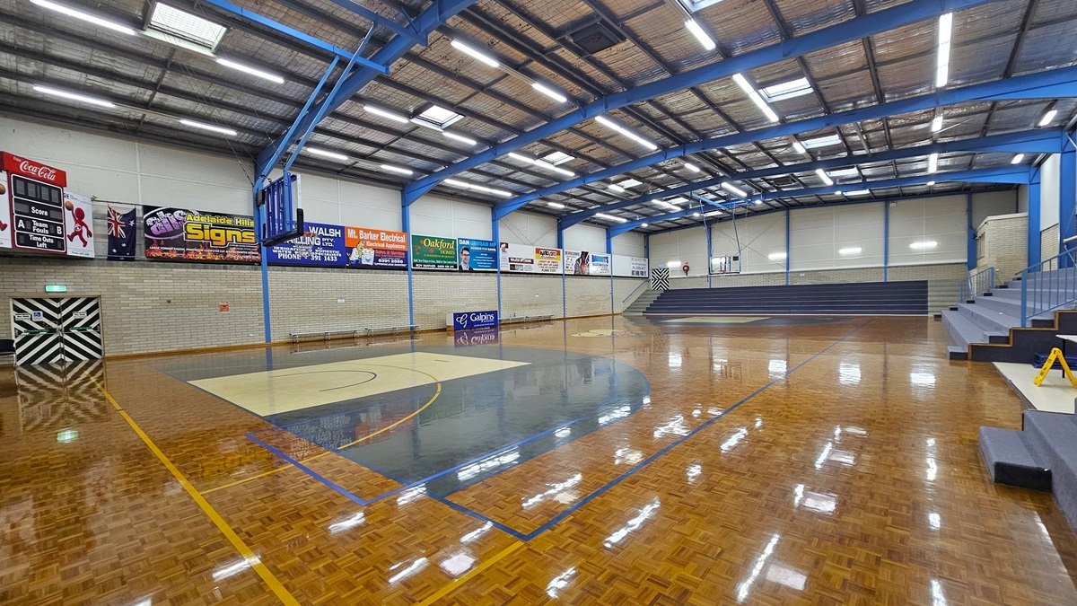Court 1 has a polished timber parquet floor with game markings. Tiered seating on the right and middle provides spectator views. Walls around the courts display advertising materials. The ceiling features exposed steel beams, fluorescent lights, and insulation foams. A digital screen is in the left corner, and below is an emergency exit door. The basketball boards are placed on both sides.
