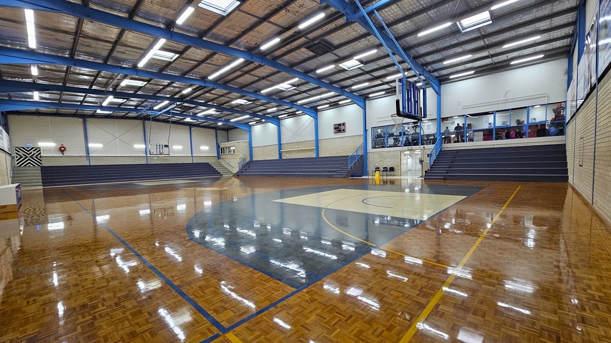 Court 1 features a polished timber parquet floor with game markings. Tiered seating on the left and middle provides spectator views. Additional space for spectators is located on the second level. Walls are made of white bricks. The ceiling features exposed steel beams, fluorescent lights, and insulation foams. On the left corner is an emergency exit door. Basketball boards are placed on both sides.