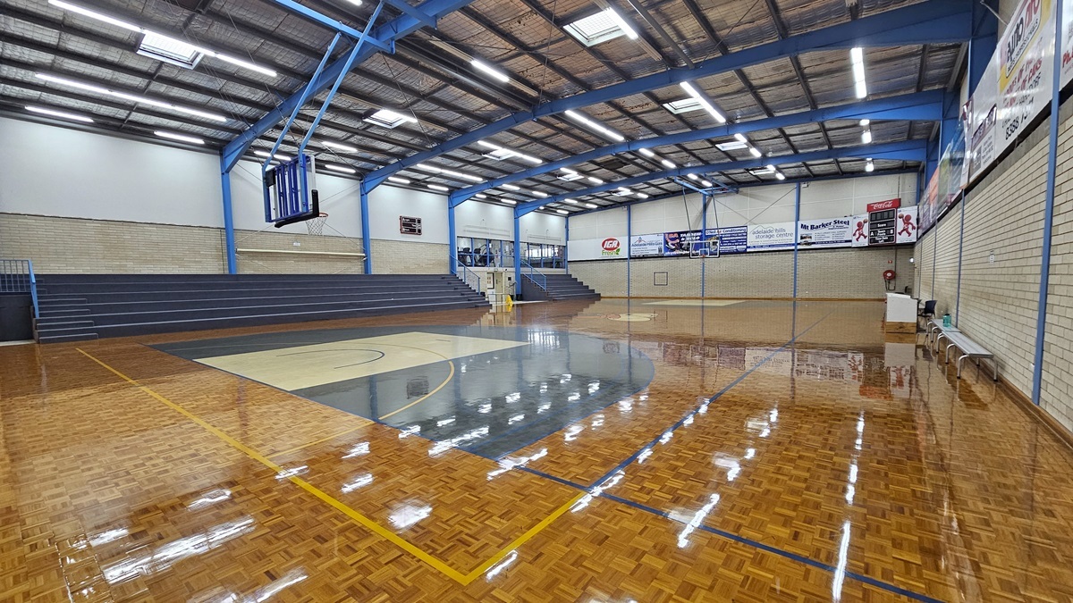 Court 1 features a polished timber parquet floor with game markings. Tiered seating is on the left, and additional space for spectators is on the second level in the left corner. Additional seats are against the right wall. A digital screen is affixed to the corner wall. Advertising posters adorn the right wall made of white bricks. The ceiling features exposed steel beams, fluorescent lights, and insulation foams. On the left corner is an emergency exit door.