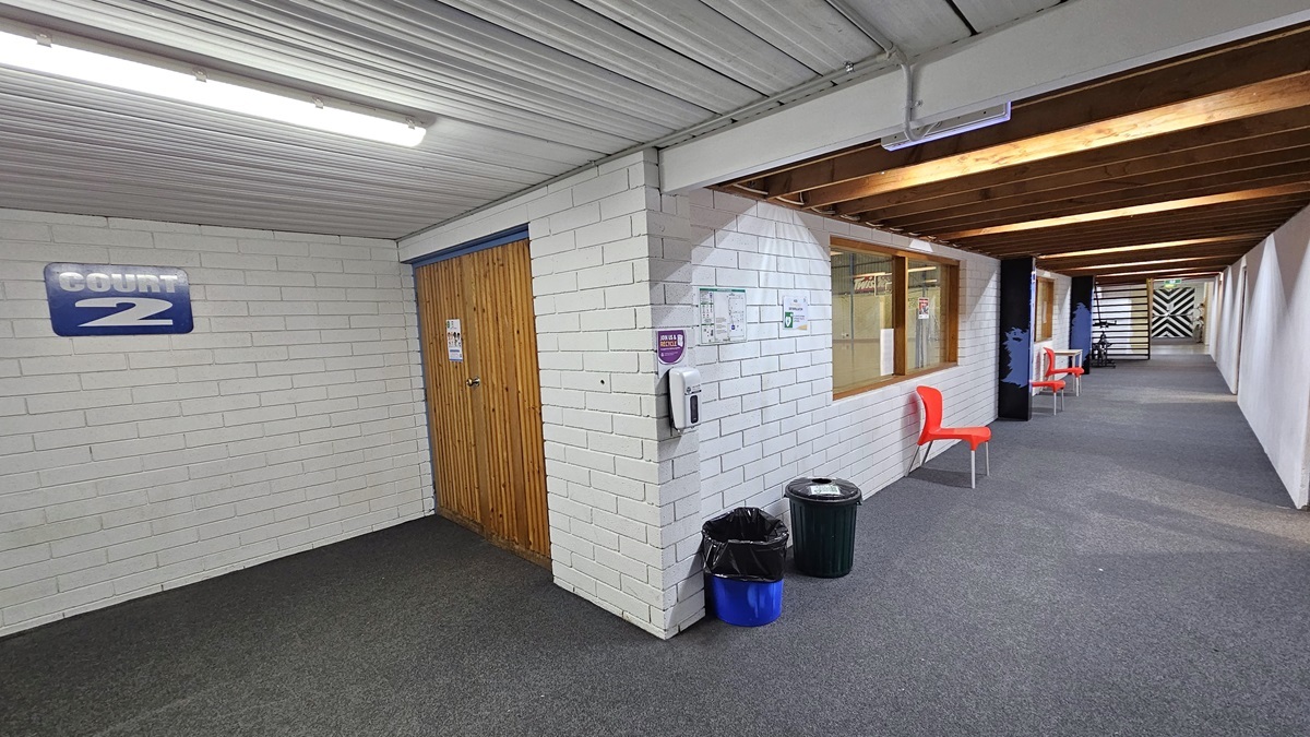 Access to Court #2 is through a door with a wooden design. The left white brick wall displays the sign "Court 2." On the right is a lobby with a wooden plank ceiling. Orange plastic chairs are against the middle wall. A hand sanitizer dispenser and two rubbish bins are near the door. The white ceiling has a corrugated design, and the floor is carpeted in grey.
