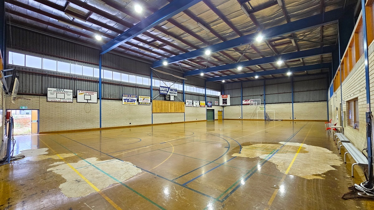 Court 2 features a sturdy concrete floor with game markings, showing signs of worn-out paint in some areas. On the left, bench chairs are available for seating. The court is enclosed by a white brick wall with boards and posters on the right. The ceiling is equipped with exposed steel beams, fluorescent lights, and insulation foams. Basketball boards are placed on both sides.
