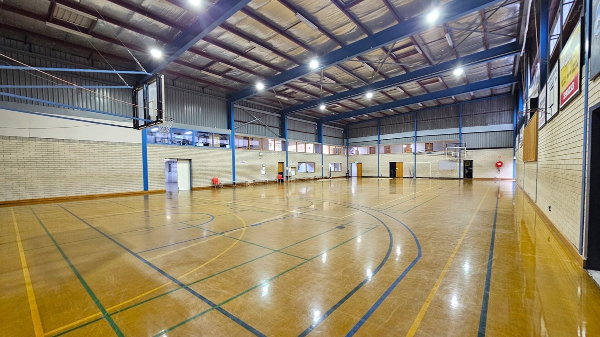 Court 2 features a sturdy concrete floor with game markings, showing signs of worn-out paint in some areas. On the left, bench chairs are available for seating. The court is enclosed by a white brick wall with boards and posters on the right. The ceiling is equipped with exposed steel beams, fluorescent lights, and insulation foams. Basketball boards are placed on both sides.