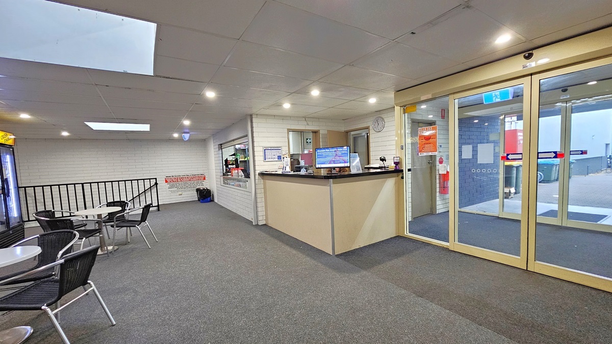 On the right is the automatic glass sliding door. Adjacent to it is a white reception table equipped with a flat-screen and reception accessories. A clock is affixed to the wall. To the right, movable chairs and tables are positioned. Next to the reception desk is a cafe. The wall is made of all-white bricks, the ceiling is white, and the floor is grey. A stair with a railing is visible at the end.