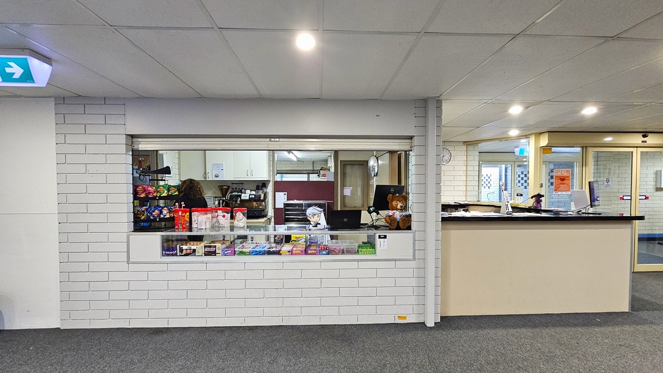 On the right is a white reception desk and an automatic glass door. In the middle is the cafe offering hot and cold drinks, desserts, and treats arranged on the display rack and on the counter. A computer, a POS machine, and a brown teddy bear are placed on the corner of the counter. The wall has a white brick design, the ceiling has a white porous design, and the floor is grey. A sign showing direction is placed on the ceiling.
