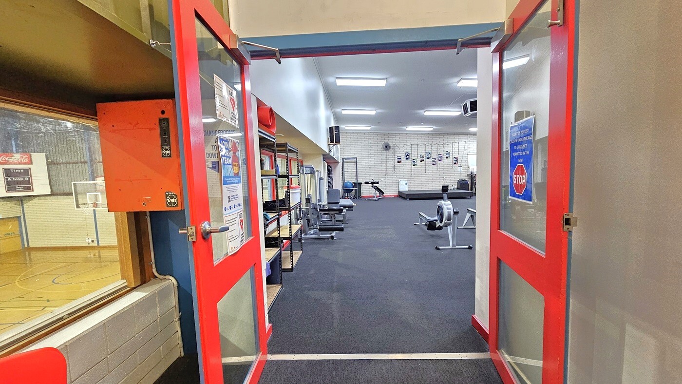 The Gymnasium is accessed through outward-opening glass doors with orange frames, adorned with affixed posters. To the left, shelves are provided for belongings. Inside, various machines are arranged, with a glass window on the left serving as a divider from the basketball court. The floor features dark grey rubber tiles, and the all-white ceiling is illuminated by aligned fluorescent lights.