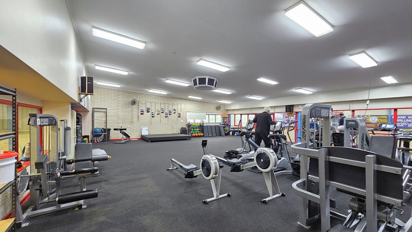 Inside the gym, machines are arranged on the right, with a person using a treadmill. Leg adductor machines are positioned on the left, alongside other machines including additional treadmills. Towards the far end, there's a small platform and a rack displaying various weights and gym accessories. The floor is made of dark grey rubber tiles, and the ceiling is adorned with white fluorescent lights.