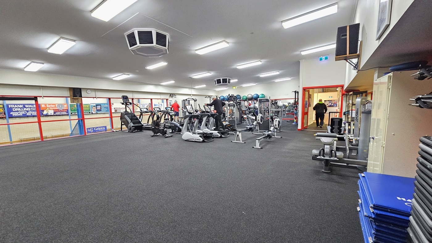 To the right is a floor-to-ceiling glass, adorned with colourful posters affixed to the wall. Cardio machines, including treadmills and step mills, are neatly lined up in the middle. Adjacent to them, adjustable benches and leg adductor machines are positioned on the left. Neatly piled exercise mats are to the left, while further along, weight equipment is stacked on a shelf, including stability balls. The floor is covered in grey rubber tiles.