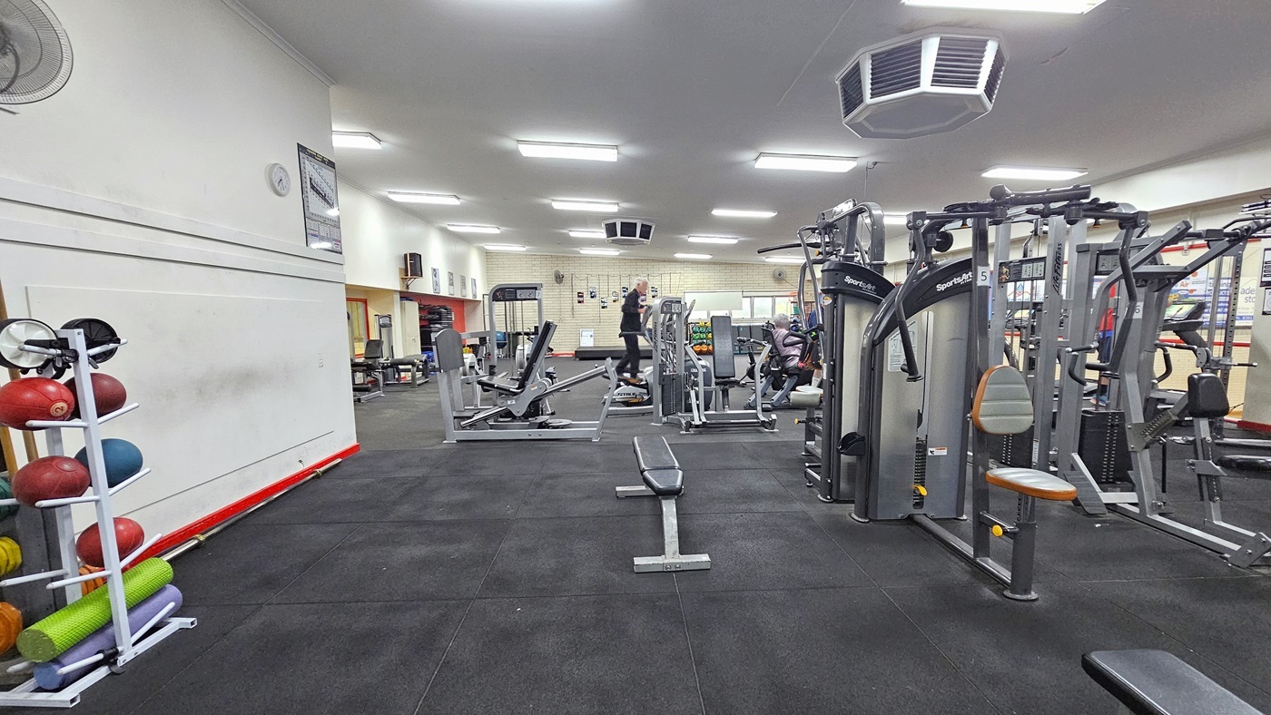 To the left is a display rack featuring medicine balls, weights, and rubber mats. A wall fan, clock, and poster adorn the top wall. On the right, various machines such as bench machines, Lat pull-ups, and treadmills are present. Two people are using the equipment. The floor is rubber tiles, and the white ceiling has rows of fluorescent lights with an octagon-shaped air conditioning unit mounted.