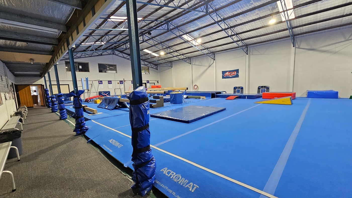 The gymnastics hall is a large industrial space featuring skylights and fluorescent lighting. The ceiling is covered with insulation foams and metal beams. White walls complement the bright blue hard foam floor. Gymnastics equipment, including mats, balance beams, vaults, and bars, is strategically arranged. On the left, there is a carpeted area with bench chairs aligned against the wall. The bottom of the posts is wrapped with foams for safety.
