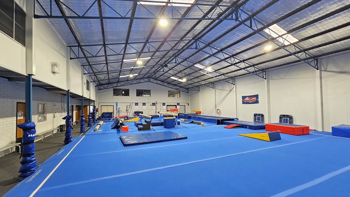 The gymnastics hall is a large industrial space featuring skylights and fluorescent lighting. The ceiling is covered with insulation foams and metal beams. White walls complement the bright blue hard foam floor. Gymnastics equipment, including mats, balance beams, vaults, and bars, is strategically arranged. On the left, there is a carpeted area with bench chairs aligned against the wall. The bottom of the posts is wrapped with foams for safety.