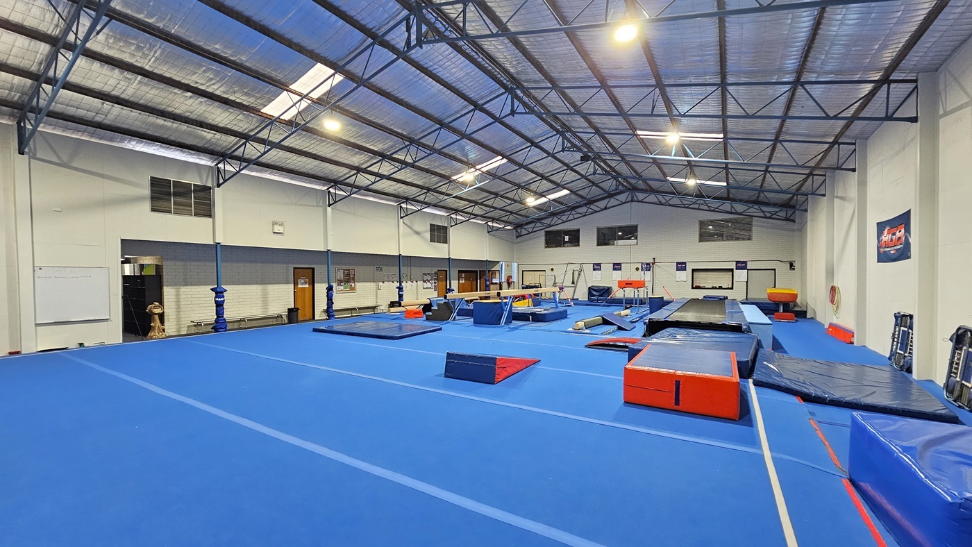 The gymnastics hall is a large industrial space featuring skylights and fluorescent lighting. The ceiling is covered with insulation foams and metal beams. White walls complement the bright blue hard foam floor. Gymnastics equipment, including mats, balance beams, vaults, and bars, is strategically arranged. On the left, there is a carpeted area with bench chairs aligned against the wall. The bottom of the posts is wrapped with foams for safety. A banner is affixed to the right wall.