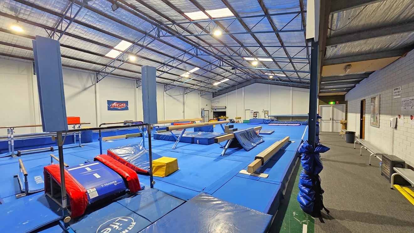 The gymnastics hall is a spacious industrial area with skylights and fluorescent lighting. The ceiling is covered with insulation foams and metal beams. White walls complement the bright blue hard foam floor. Gymnastics equipment, such as mats, balance beams, vaults, and bars, is strategically arranged. On the right, there is a carpeted area with bench chairs aligned against the wall. Posters and sticker reminders are affixed to the wall. The bottom of the posts is wrapped with foam for safety.