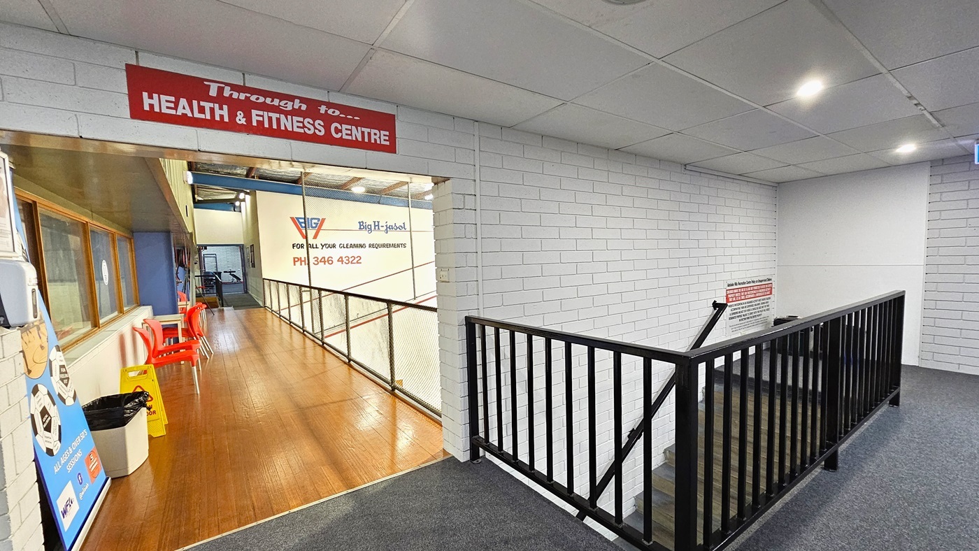 Ahead is the viewing platform for the Squash Courts, featuring a timber wood floor and a secure metal railing with a net. Red plastic chairs are positioned against the wall for seating. On the left, glass windows, a large banner, a wall-mounted hand sanitizer, a rubbish bin, and a safety sign are visible. To the right, stairs with metal railings lead down. Signages and reminders are painted on the brick walls.