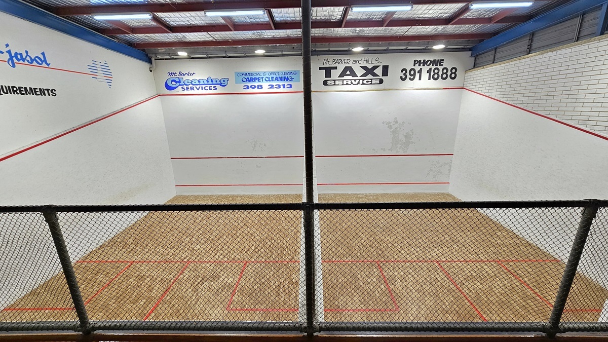 The Squash Court is visible from the viewing deck, enclosed by metal railings with a net. The court features a timber floor marked with vibrant red lines. Advertisements adorn the white walls, and the high ceiling is illuminated by fluorescent lights and equipped with insulation forms.
