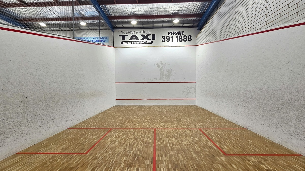 The Squash Court is visible from the viewing deck, enclosed by metal railings with a net. The court features a timber floor marked with vibrant red lines. Advertisements adorn the white walls, and the high ceiling is illuminated by fluorescent lights and equipped with insulation forms.